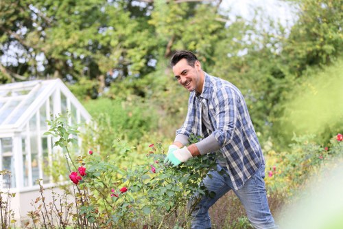 A man with dark hair and a light complexion is tending to a flowering garden, wearing a blue and white checked shirt, jeans, and gardening gloves. He is smiling and appears to be pruning or caring for the vibrant rose bushes, which display pink and red blossoms. The garden layout includes a well-maintained lawn with lush green grass, a variety of shrubs, and flower beds bordering a paved pathway. In the background, there is a glass greenhouse with white framing, set amidst trees and naturally landscaped greenery under a bright, clear sky, suggesting a warm, sunny day. The outdoor space is neat and organized, combining cultivated flower beds, mature trees, and lawn areas suitable for maintenance and landscaping services. The scene illustrates professional gardening activity, aligned with local lawn and garden care offerings, with a focus on natural tones, plant diversity, and garden structure typical of high-quality outdoor landscaping in a residential setting, possibly in Highbury area.